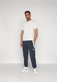 T-shirt branca de gola redonda combinada com calças de treino azuis marinho com detalhes brancos e emblemas de logotipo, complementadas por ténis desportivos brancos.