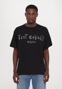 Melnas kokvilnas T-krekls ar īsām piedurknēm, ar baltu grafisku tekstu "Just Cavalli Milano" priekšpusē. Ikdienišķs piegriezums ar apaļu izgriezumu.