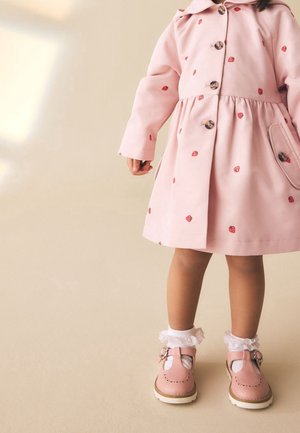 Enfant portant un manteau rose avec des imprimés fraises, des chaussettes blanches à volants et des chaussures roses à boucle, debout sur un fond beige.
