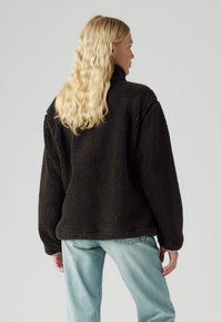 Chaqueta de forro polar negra con cuello alto, ajuste holgado, tela texturizada y mangas anchas, combinada con jeans de mezclilla claros.