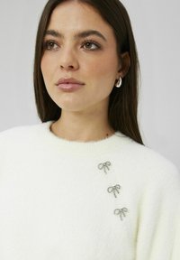 Pull blanc duveteux avec trois nœuds décoratifs argentés sur l'épaule. Encolure ronde et manches raglan. Texture douce.