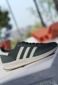 Baskets de sport vert foncé et blanc avec trois bandes blanches et semelle en gomme, posées sur une surface carrelée avec un arrière-plan extérieur flou.