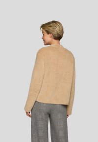 Beige, flauschige Jacke mit rundem Ausschnitt und langen Ärmeln, kombiniert mit grauen, karierten Hosen. Weiche Textur und entspannte Passform.