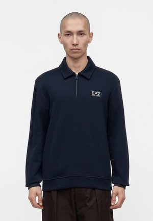 Jeune homme portant un pull marine à demi-fermeture éclair avec un petit logo EA7 Emporio Armani sur la poitrine, debout devant un fond blanc uni.
