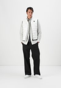 White quilted puffer vest with black zippers, worn over a light gray button-up shirt and black trousers. Model stands on a plain background.