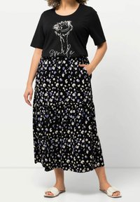 Nainen, jolla on päällään musta t-paita, jossa on hymyilevän strutsin kuvasto, sekä mustat hameet, joissa on valkoisia ja violetteja abstrakteja pilkkuja.