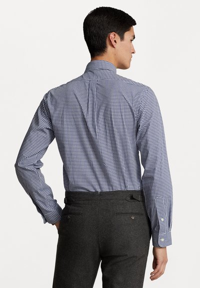 Knopenhemd met een marineblauw en wit ruitpatroon, lange mouwen en een staande kraag. Draagt met grijze broeken met achterzakken.