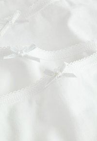 Witte katoenen cami met delicate kanten randen en kleine satijnen strikjes op de bandjes. Zachte textuur met een gladde, gelijkmatige afwerking.