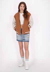 Giacca varsity in corduroy marrone e pile crema, top bianco corto, gonna in denim strappata, sneakers bianche, calze a strisce verdi e bianche.