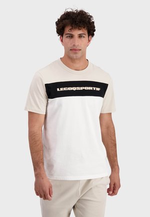 Camiseta de hombre con un diseño en bloques de color crema y blanco, con una franja horizontal negra y "LECOQSPORTIF" impreso en beige.