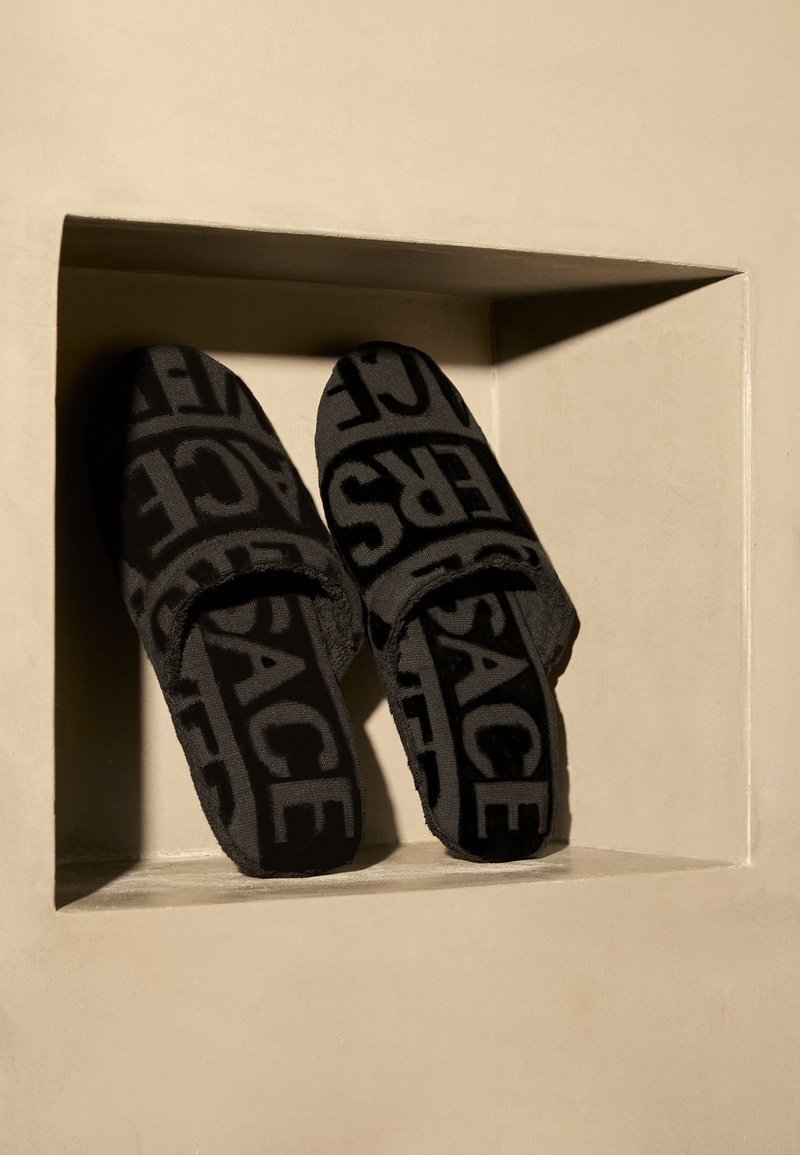 Sorte plyssandaler med en struktureret overflade, der har et gentaget logomønster. Åben tå-design præsenteret i en nedsænket udstilling.