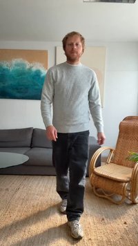 Suéter cinza de malha com um padrão texturizado, jeans escuros e ténis claros. O fundo apresenta um sofá cinza, uma mesa de vidro e uma cadeira de vime.