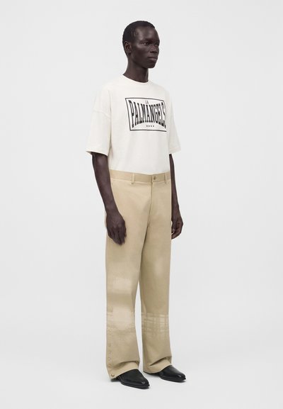 Άνδρας που φοράει oversized off-white μπλούζα με μαύρο κείμενο και μπεζ παντελόνι με φαρδιά μπατζάκια και κουμπιά στις μανσέτες, στέκεται μπροστά σε απλό φόντο.