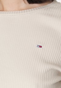 Beige ribbat tröja med bred halsringning och en liten broderad logotyp med blå, vit och röd rand på vänster sida.