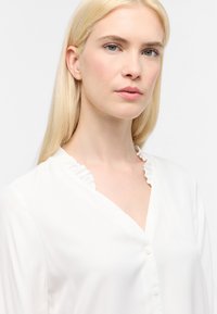 Weiße Bluse mit Rüschensaum, Knopfverschluss und glattem Stoff. Verfügt über lange Ärmel und ein V-Ausschnitt-Design.