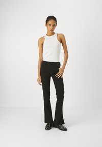 Pantalones de campana negros de algodón, combinados con una blusa sin mangas blanca. Los pantalones presentan un diseño clásico de cinco bolsillos y un cierre de botón.