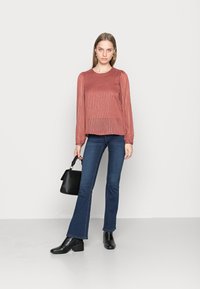Blusa de cor ferrugem com riscas verticais, mangas longas e bufantes, combinada com jeans flare azul escuro e botins pretos. Inclui uma mala preta.