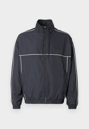 Veste coupe-vent bleu marine avec col haut, détails de passepoil blanc le long des épaules, poignets élastiques et poches avant zippées.