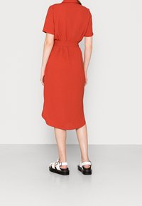 Abito rosso a maniche corte con cintura in vita e lunghezza midi. Presenta una texture liscia e un orlo curvo e sottile. Indossato con sandali bianchi.