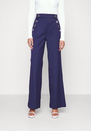 Donkerblauwe hoge taille broek met wijde pijpen en zes decoratieve knopen, gecombineerd met een witte geribbelde top met lange mouwen en witte open-teen hakken.