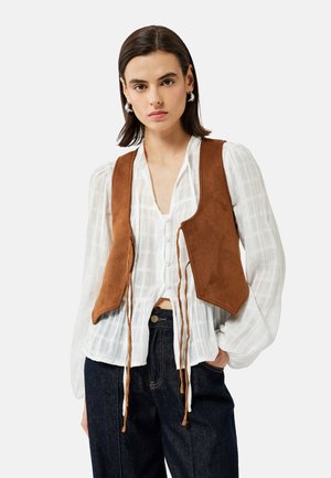 Brauner Wildledervest mit figurbetonter Passform, vorne mit Bindebändern, getragen über einer weißen, strukturierten Bluse mit Knopfverschluss und Puffärmeln.