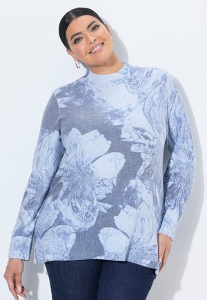 Kvinde smiler iført en lyseblå blomstret langærmet sweater og mørke jeans mod en ensfarvet lys baggrund.