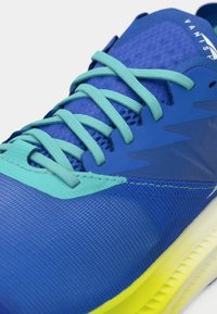Zapatilla deportiva azul con material de malla, que presenta cordones y detalles en turquesa, una parte superior texturizada y una suela degradada que pasa a amarillo.