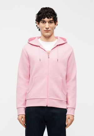 Mladý muž s kučeravými vlasmi, oblečený v svetloružovej mikine na zips a tmavých nohaviciach, stojaci pred jednoduchým svetlým pozadím.