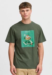 Camiseta de algodón verde oliva con una ilustración gráfica centrada de un postre, con texto arriba y abajo. Mangas cortas, corte relajado.