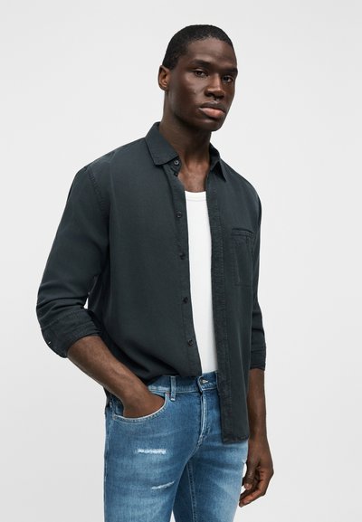 Chemise boutonnée vert foncé avec une poche poitrine, portée sur un débardeur blanc. Associée à un jean bleu clair déchiré.