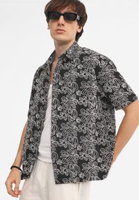 Camicia nera a maniche corte con ricamo floreale bianco, tessuto leggero, design con colletto e vestibilità rilassata. Maglietta bianca sotto visibile.