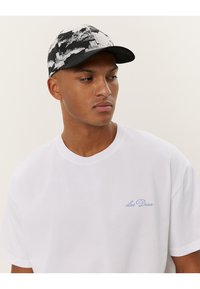 Schwarz-weiß gemusterte Kappe mit gebogenem Schirm, getragen von einem Modell in einem schlichten weißen T-Shirt mit blauem gesticktem Text.