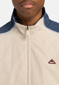 Light beige zip-up jacket with a high collar, featuring dark blue shoulder accents and a red triangular logo on the left side.