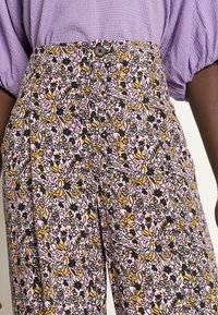 Calças florais de cintura alta em rosa, preto e amarelo. Apresentam um fecho de botão, padrões de flores intrincados e uma textura de tecido macio.