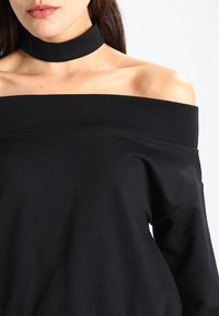 Žena oblečená v černém topu bez ramínek a odpovídajícím černém náhrdelníku, ukazující dekolt a levé rameno na bílém pozadí.