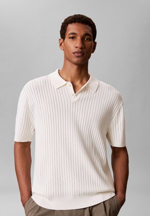 Mężczyzna stoi z rękami w kieszeniach, ubrany w białą, krótką koszulkę dzianą z kołnierzykiem oraz beżowe spodnie na jednolitym szarym tle.