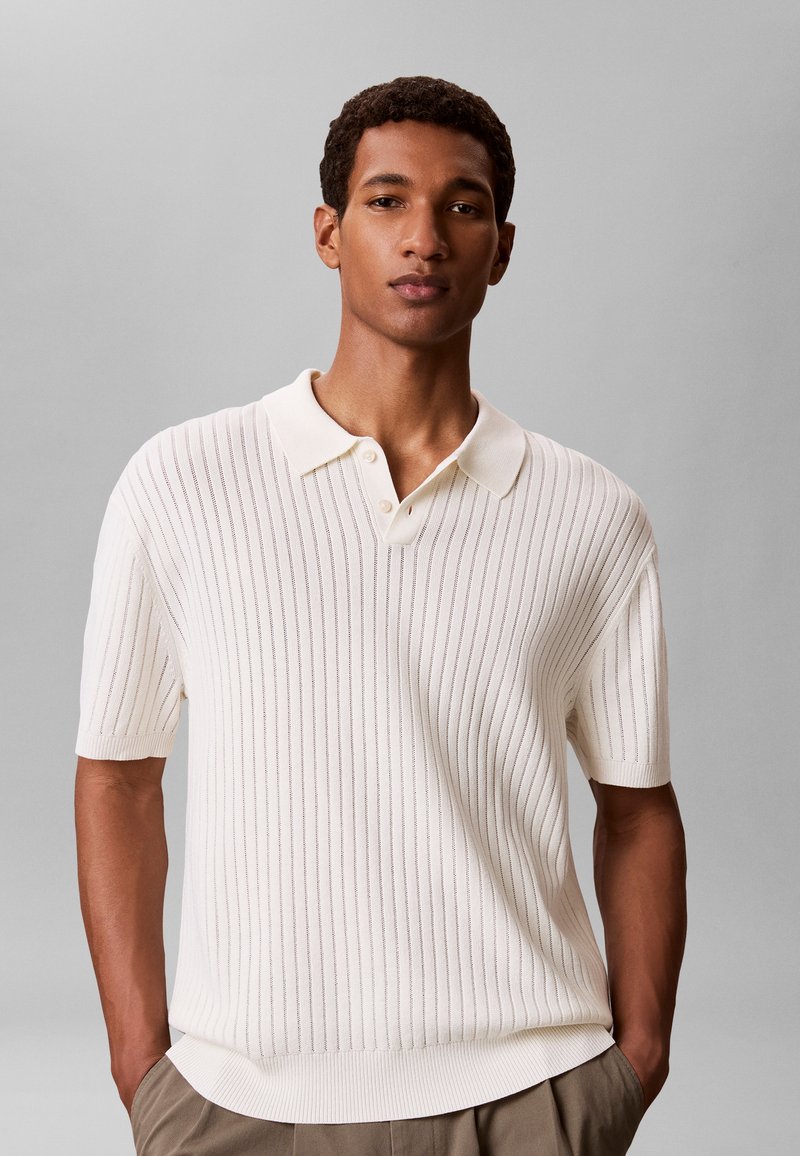 Homme debout les mains dans les poches, portant une chemise en maille blanche à manches courtes avec col et un pantalon beige sur un fond gris uni.