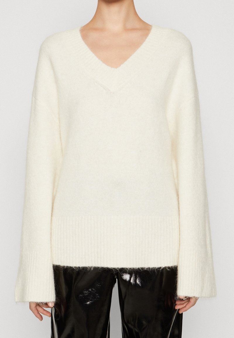 Pull à col en V blanc crème et duveteux à manches longues, associé à un pantalon en vinyle noir brillant, porté par une personne sur un fond uni.