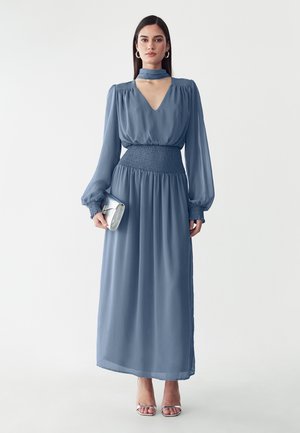 Mujer con vestido largo azul con mangas abullonadas y cintura fruncida, sostiene un bolso de mano plateado y lleva tacones plateados abiertos en un fondo blanco.