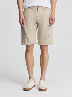 Beżowe spodenki do kolan z podwiniętymi brzegami i przetarciami, noszone z białymi trampkami i białą koszulką.