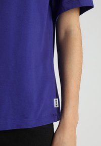 Osoba ubrana w fioletową koszulę z krótkim rękawem, z małą białą metką "NSW" blisko końca po prawej stronie, na jednolitym tle.