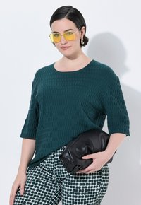 Teal strikket sweater med tekstur og korte ærmer, parret med mønstrede grønne og hvide bukser. Holder en sort læder clutch. Gule solbriller.