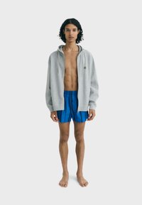 Grijze zip-up hoodie met een voorkeurszak, gedragen over blauwgestreepte zwembroek. Beschikt over lange mouwen en een relaxte pasvorm. Blootvoets.