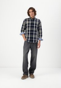 Marineblaues und weißes Karomuster-Hemd mit umgeschlagenen hellblauen Ärmeln, kombiniert mit grauen Jeans und braunen Schuhen. Das Model steht in einer neutralen Umgebung.