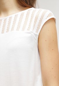 Vit kortärmad topp med texturerade meshdetaljer på axlarna. Slät tyg, rund halsringning och avslappnad passform. Minimalistisk design.