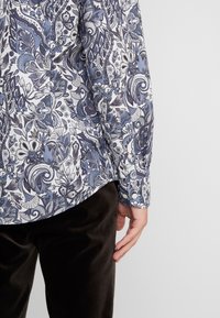 Osoba w longsleeve'ie z misternymi niebiesko-białymi wzorami paisley i ciemnymi spodniami, stojąca na jednolitym tle.