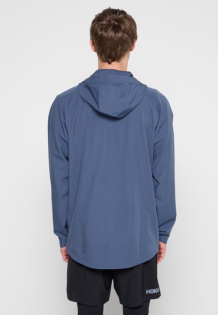 Veste à capuche bleue avec une texture lisse, dotée d'un ourlet arrière plus long et de manches raglan, portée sur des shorts noirs.