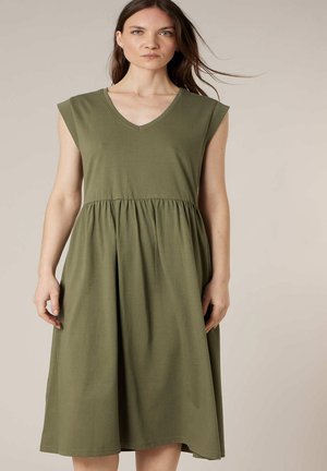 Femme aux longs cheveux bruns portant une robe midi olive verte sans manches avec un col en V et une taille froncée, debout devant un fond uni.
