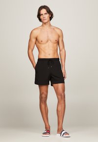 Sorte svømmedrakter med elastisk linning og snøring, med sidelommer. Kombinert med hvite sandaler med røde og blå striper.