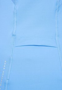 Ljusblå, elastisk sporttyg med en mjuk yta. Har synliga sömmar och en fickdetalj i mitten. Små reflexpunkter på ena sidan.